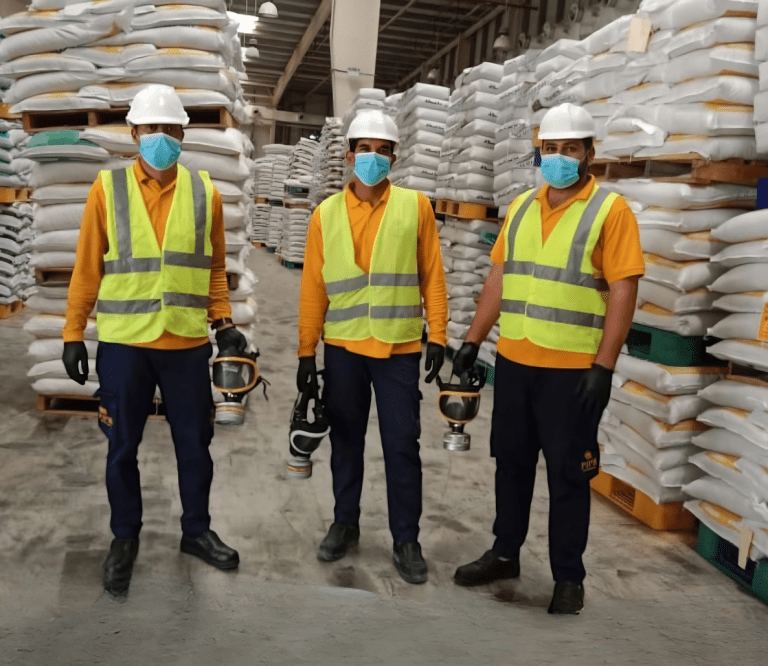 pest control Three workers in safety gear pose in a warehouse filled with stacked bags, emphasizing safety and teamwork in fumigation pest control service. PIPM